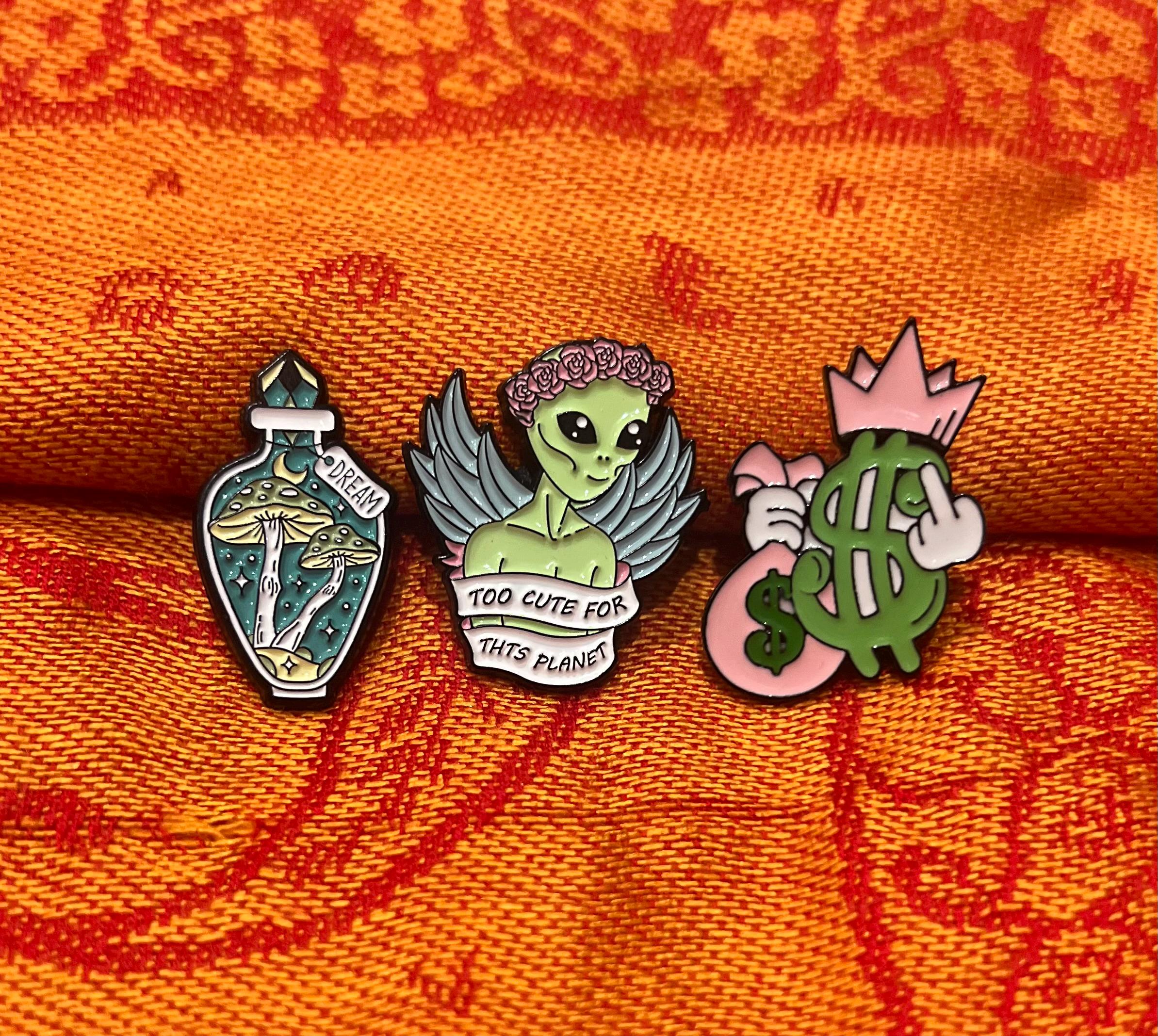 May include: Three enamel pins. One is green and features an alien with the text 'Too cute for this planet'. Another is pink and features a hand holding a dollar sign. The last is green and features a bottle with mushrooms and the text 'Dream'.