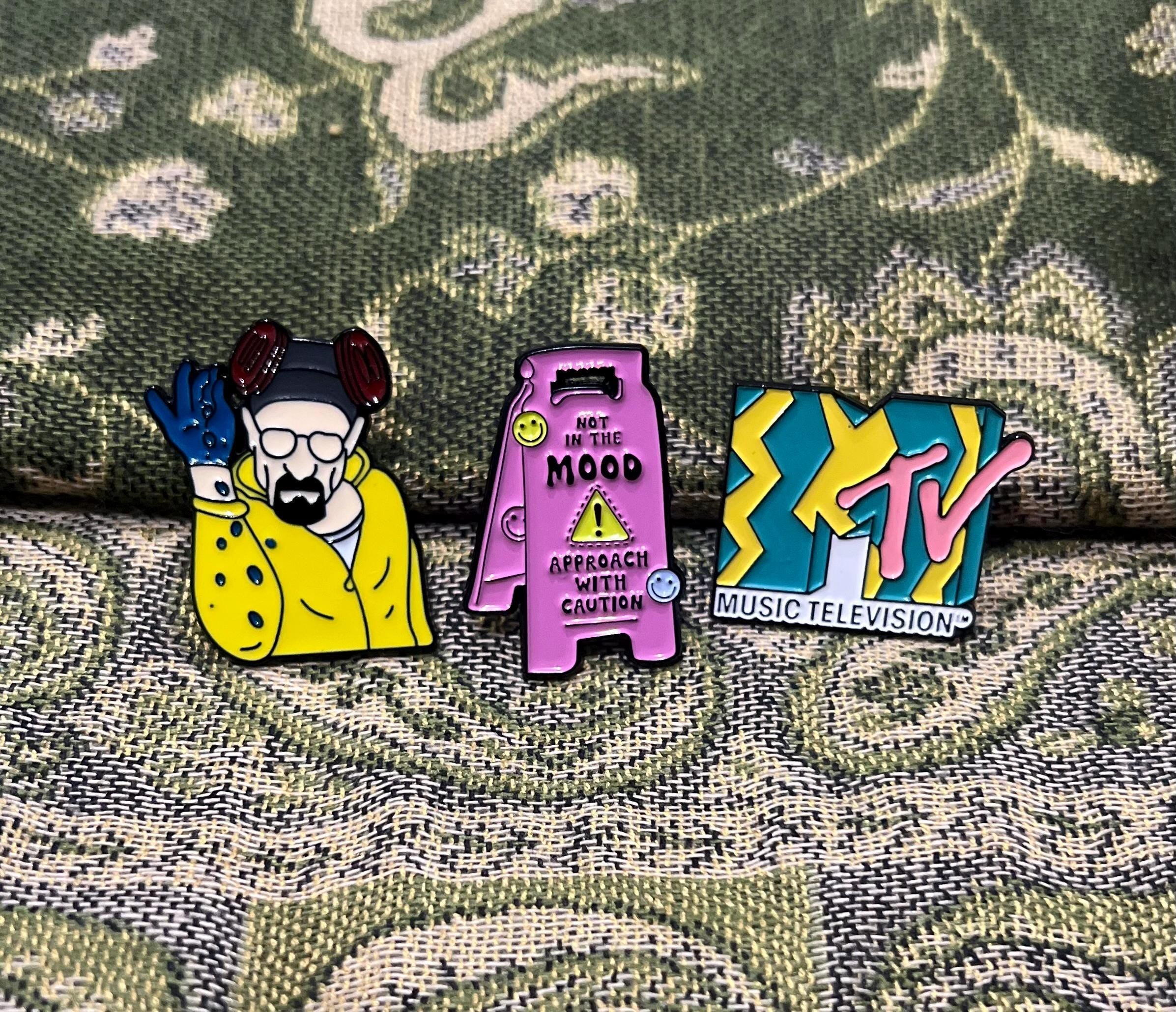 May include: Three enamel pins. One pin is pink and has a yellow caution sign with the text 'Not in the Mood Approach with Caution'. Another pin is yellow and green with the text 'MTV Music Television'. The last pin is yellow and blue and depicts a cartoon character wearing a yellow jacket and blue gloves.