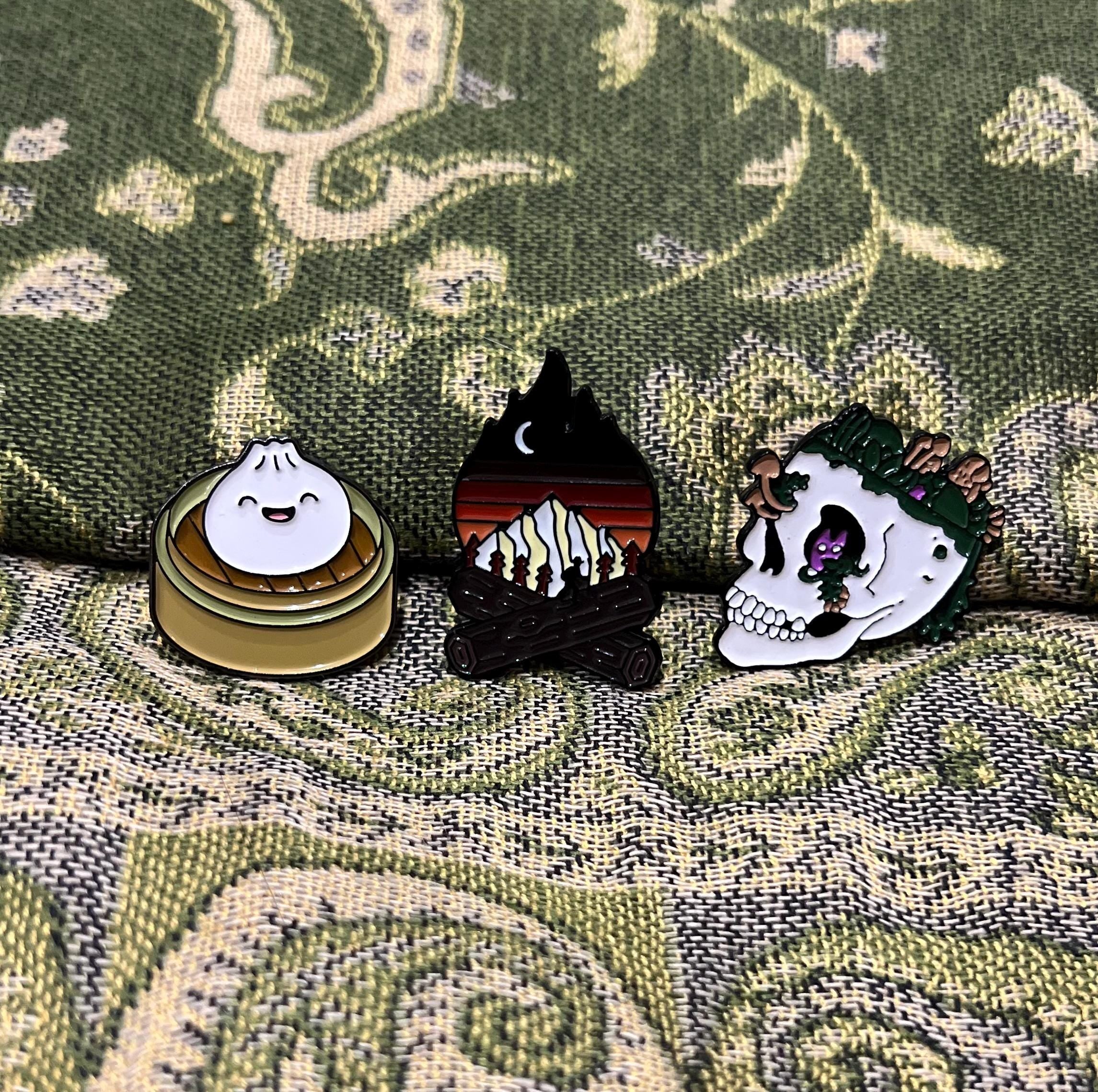 May include: Three enamel pins: a white dumpling with a smiling face in a yellow and brown steamer basket, a black campfire with a mountain range and a crescent moon behind it, and a white skull with green leaves and mushrooms growing out of the top of it.