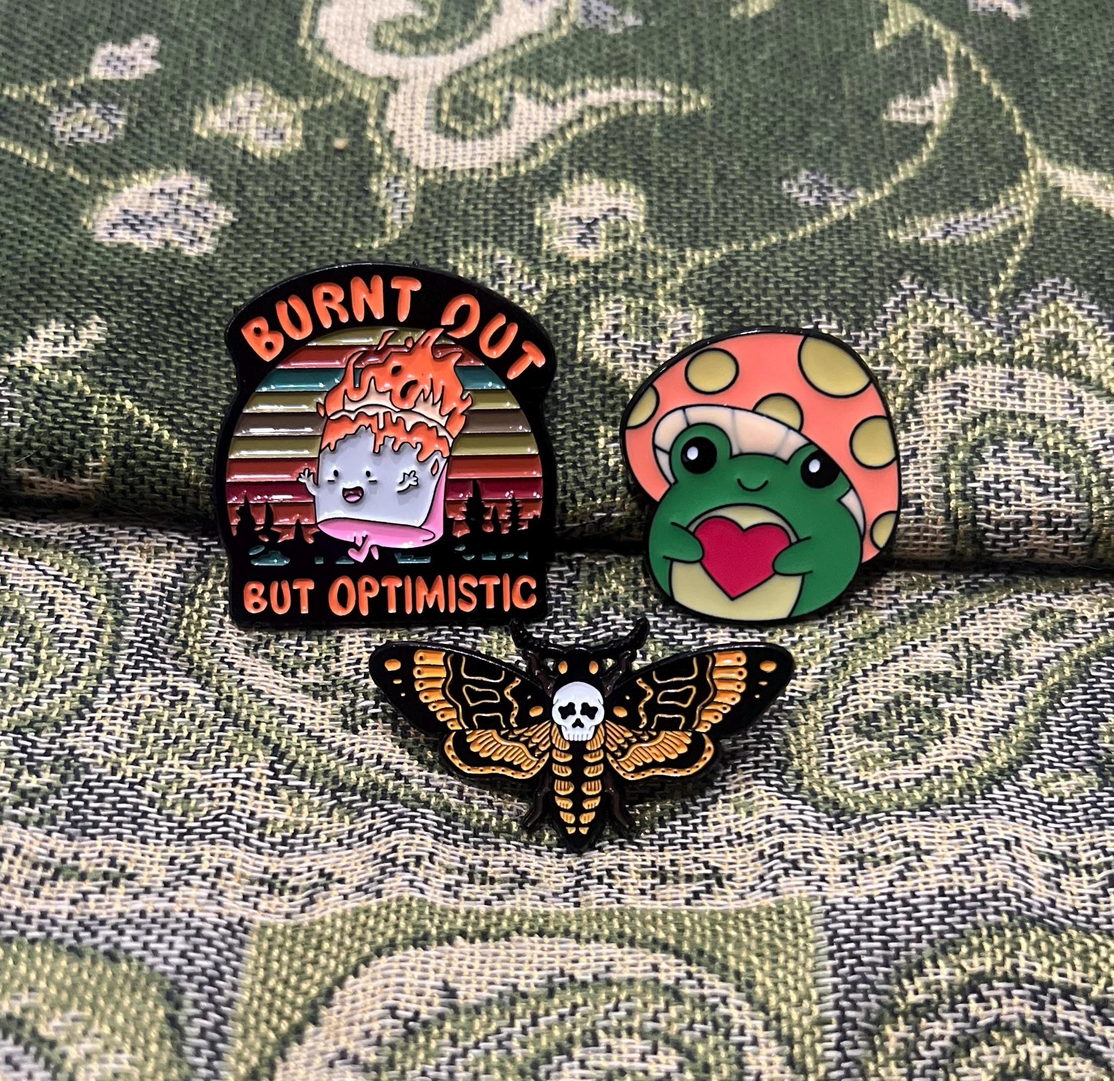May include: Three enamel pins. One pin is orange and black with the text 'Burnt Out But Optimistic'. Another pin is green and pink with a frog holding a heart. The third pin is black and yellow with a moth with a skull on its chest.
