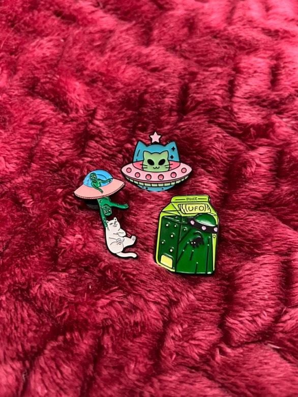 May include: Four enamel pins featuring green alien characters. One pin features a green alien in a flying saucer, another pin features a green alien in a milk carton with the word 'JUICE' and 'UFO' printed on the carton, another pin features a green alien holding a white cat, and another pin features a green alien with a star on its head in a flying saucer.