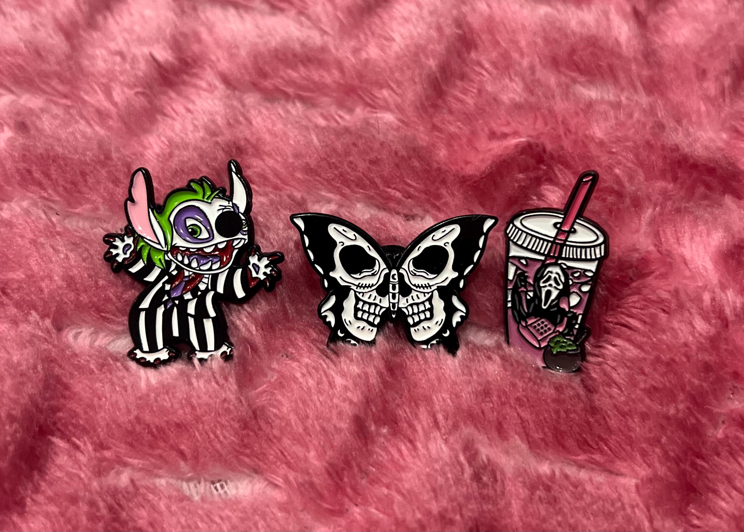 May include: Three enamel pins on a pink fuzzy surface. The pins are a black and white striped character with green ears, a black and white butterfly with two skulls, and a pink and white cup with a ghost inside.