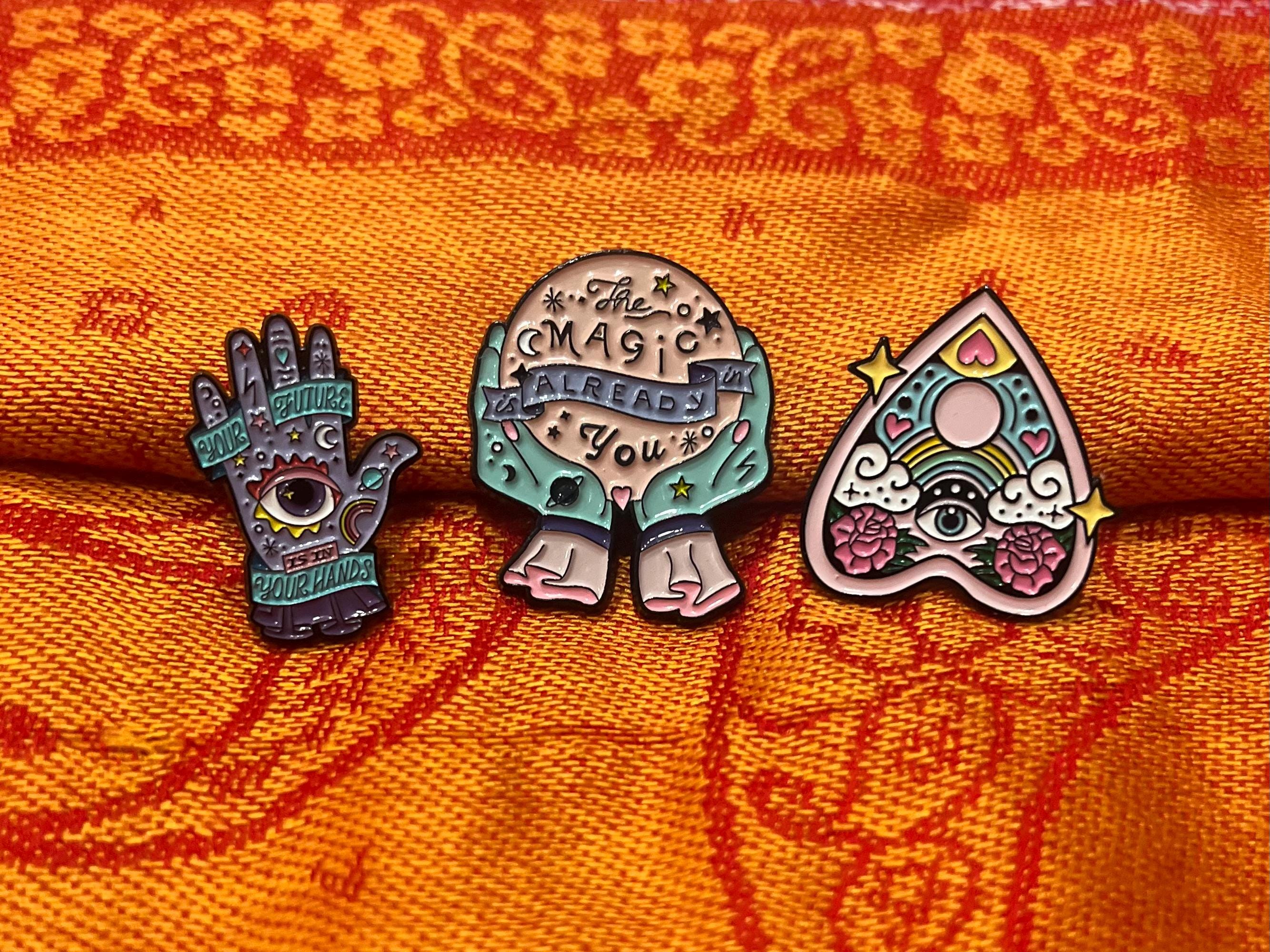 May include: Three enamel pins with a whimsical, magical theme. One pin is a hand with an eye and the text 'Your Future Is In Your Hands'. Another pin is a crystal ball with hands holding it and the text 'The Magic Is Already In You'. The third pin is a pink heart-shaped  pendant with an eye and rainbow and the text 'Love'.