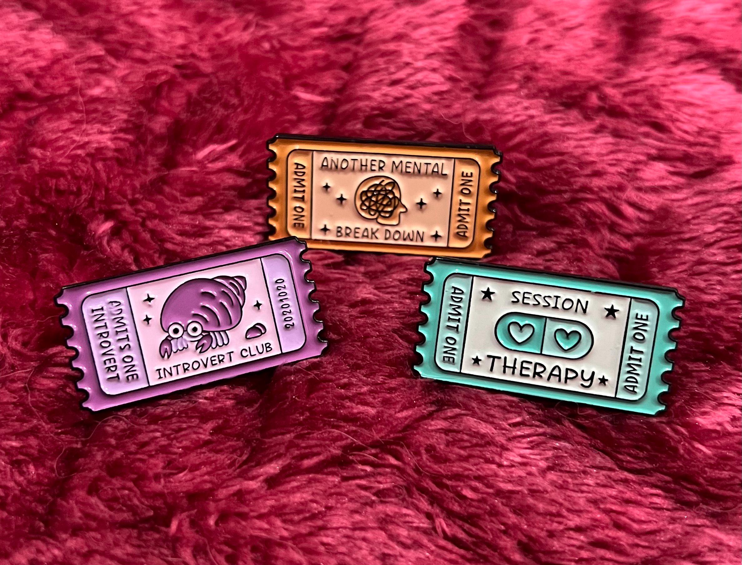 May include: Three enamel pins shaped like ticket stubs. The first pin is purple and says 'Admits One Introvert' and has a small crab illustration. The second pin is orange and says 'Another Mental Break Down' and has a brain illustration. The third pin is blue and says 'Admit One Session Therapy' and has two hearts inside a pill shape.
