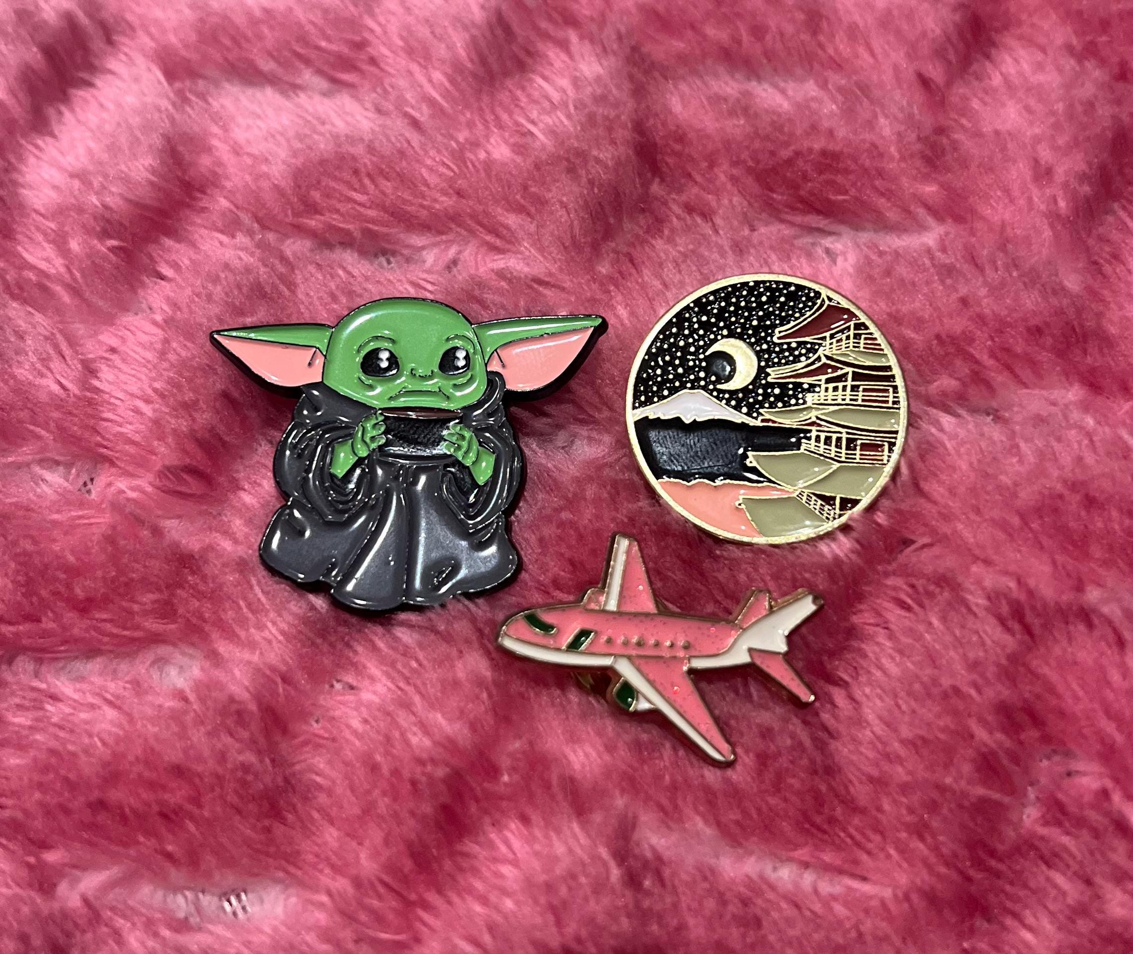 May include: Three enamel pins on a pink fuzzy surface. One pin is a green alien character with a black robe. Another pin is a pink airplane. The last pin is a gold and black pin with a mountain scene and a crescent moon.