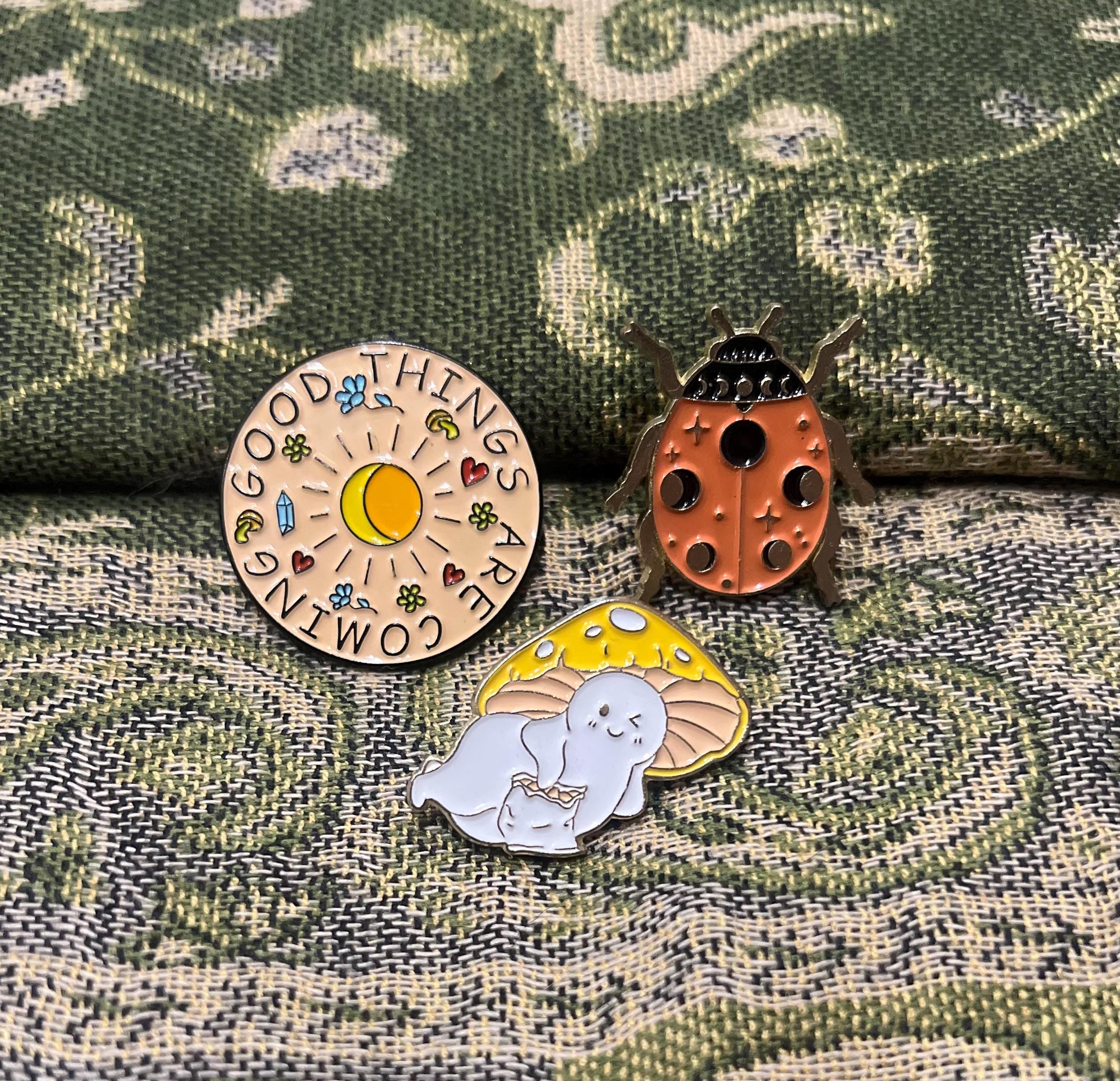 May include: Three enamel pins. One pin is round and features a sun, moon, and stars with the text 'Good Things Are Coming.' Another pin is a ladybug with a black and white pattern. The third pin is a mushroom with a white creature inside.