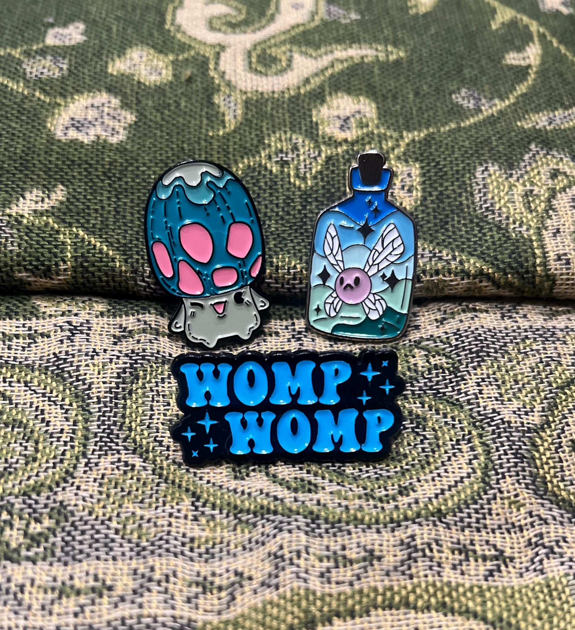 May include: Three enamel pins, one with a blue and green mushroom with pink spots, one with a blue bottle with a pink fairy inside, and one with the text 'WOMP WOMP' in blue with black outlines and silver glitter.