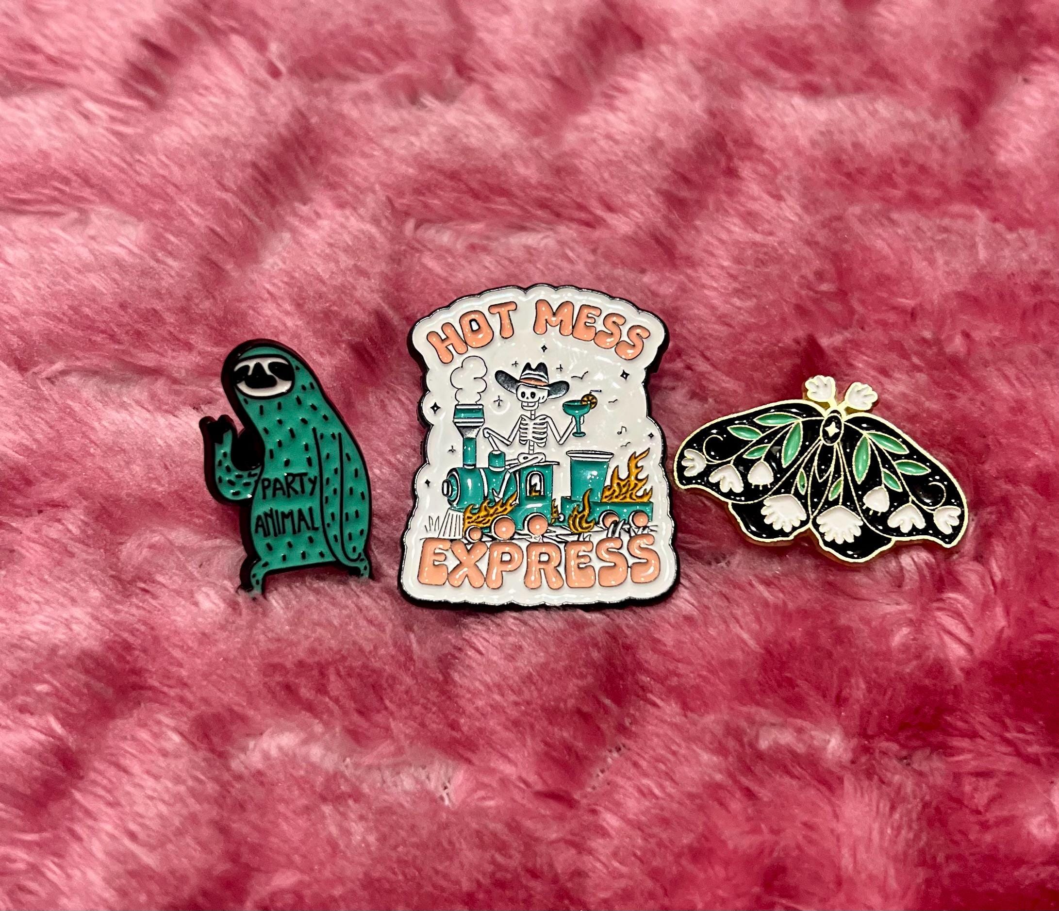 May include: Three enamel pins. A green sloth pin with the words 'Party Animal' on its belly. A black and white butterfly pin with white flowers. A white pin with a skeleton riding a train with the words 'Hot Mess Express'.