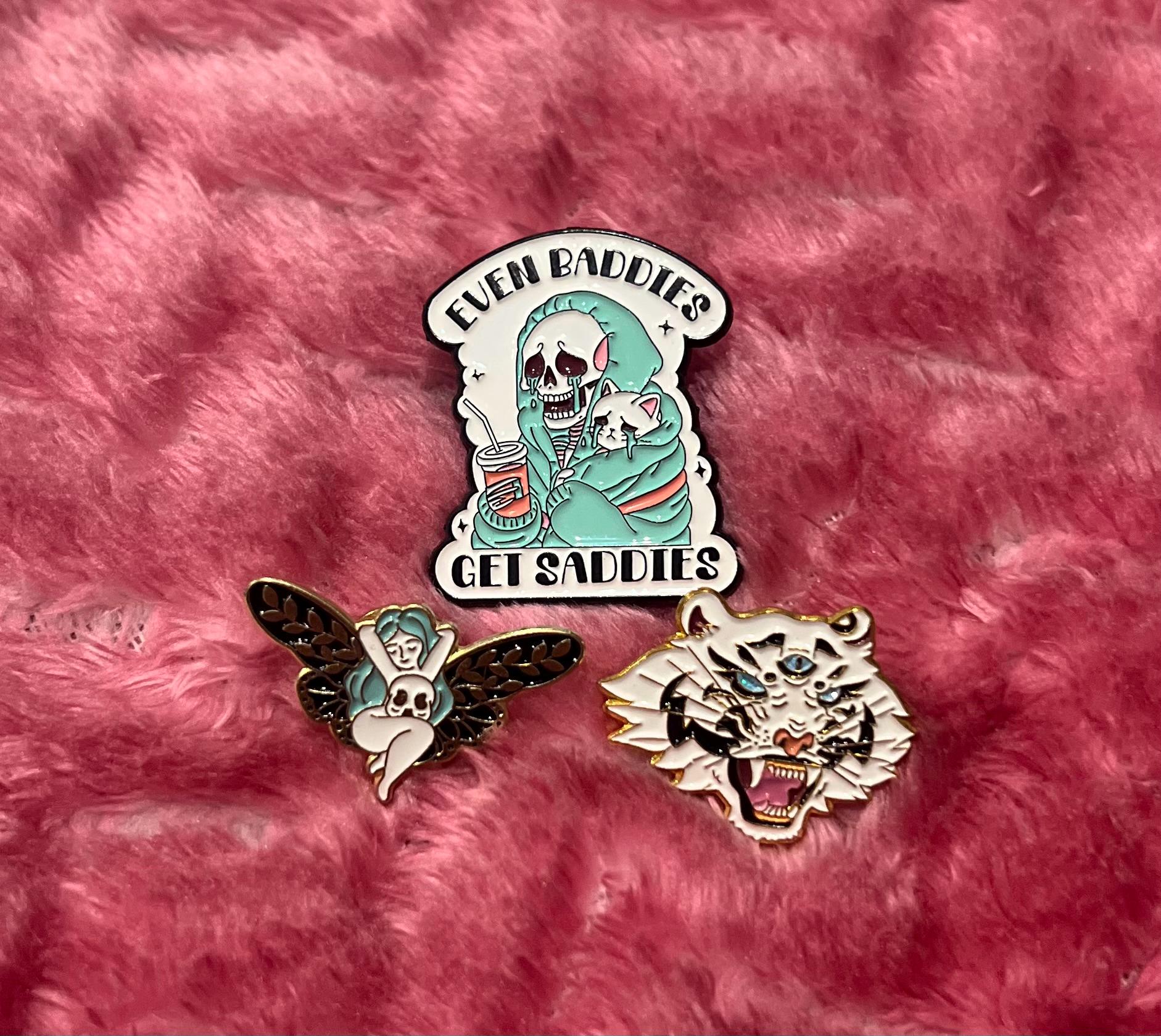 May include: Three enamel pins on a pink fuzzy surface. The first pin is a skeleton wearing a teal hooded sweatshirt holding a cup and a small cat. The text on the pin reads 'Even Baddies Get Saddies'. The second pin is a white fairy with brown wings. The third pin is a white tiger with three eyes and an open mouth.