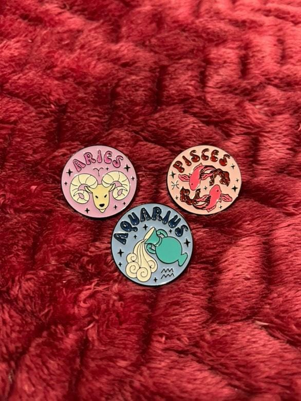 May include: Three enamel pins with zodiac signs. The pins are round and have a white background with black outlines. The first pin has a pink ram and the text 'Aries' in pink letters. The second pin has a blue water pitcher and the text 'Aquarius' in blue letters. The third pin has two orange fish and the text 'Pisces' in orange letters.