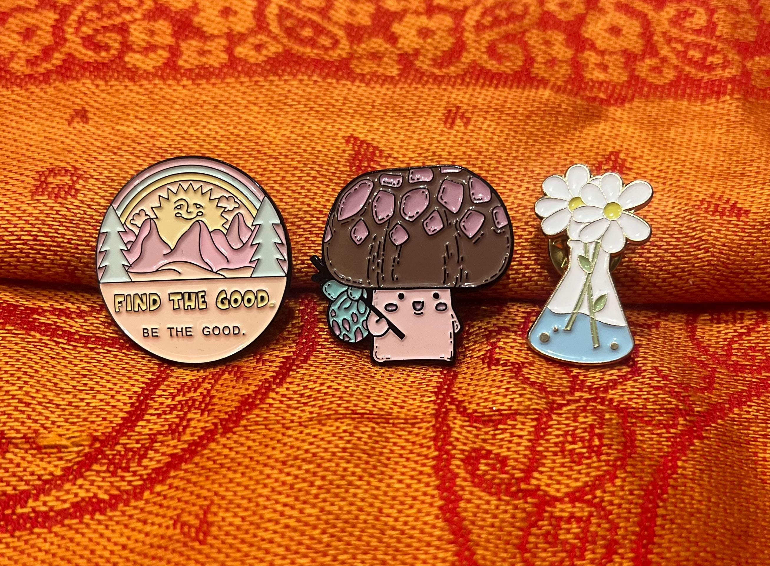 May include: Three enamel pins. One is a rainbow colored mountain scene with the text 'Find the good. Be the good.' The second is a brown mushroom with a pink face and a green leaf. The third is a white flower in a blue glass vial.