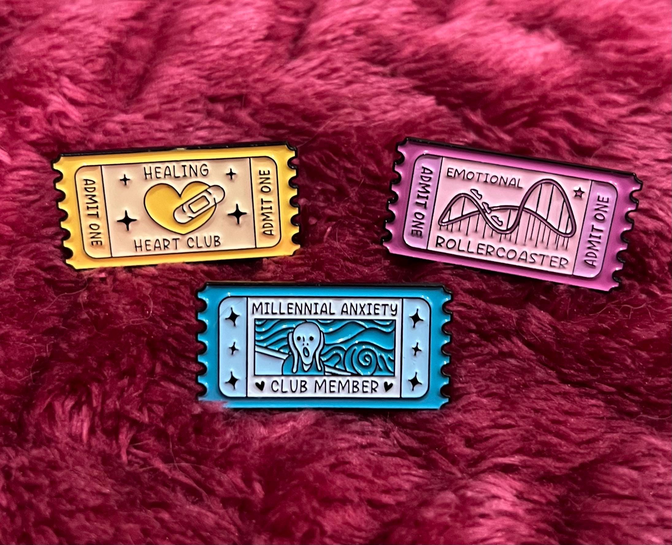 May include: Three enamel pins shaped like vintage ticket stubs. The first pin is yellow with the text 'Healing Heart Club' and a heart with a bandage. The second pin is pink with the text 'Emotional Rollercoaster' and a roller coaster. The third pin is blue with the text 'Millennial Anxiety Club Member' and a drawing of the Scream painting.