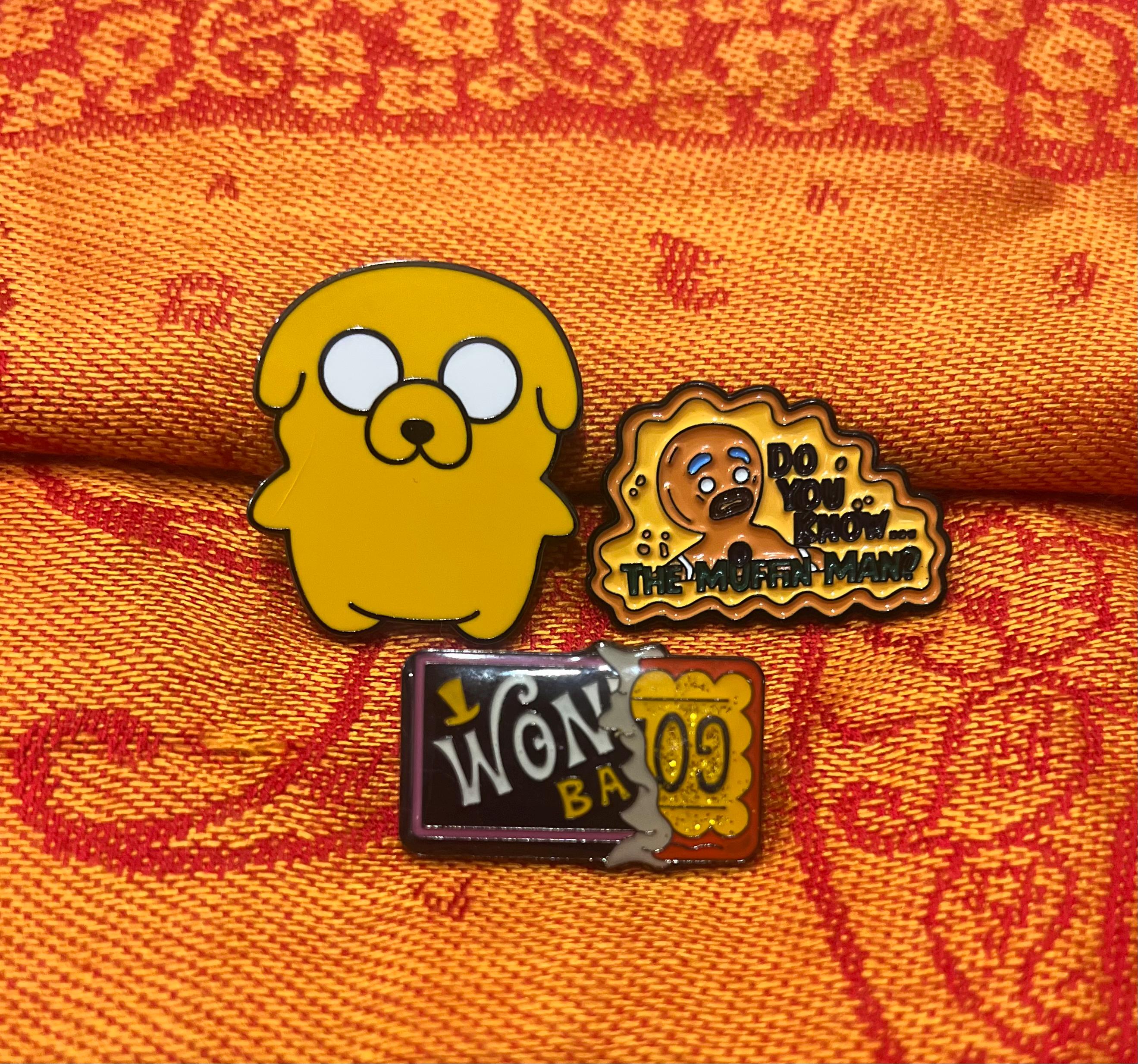 May include: Three enamel pins featuring characters from the animated television series Adventure Time. The first pin is yellow and depicts the character Finn the Human. The second pin is brown and depicts the character The Ice King. The third pin is purple and depicts the character Jake the Dog. The Ice King pin has the text 'Do you know... The Muffin Man?' on it.