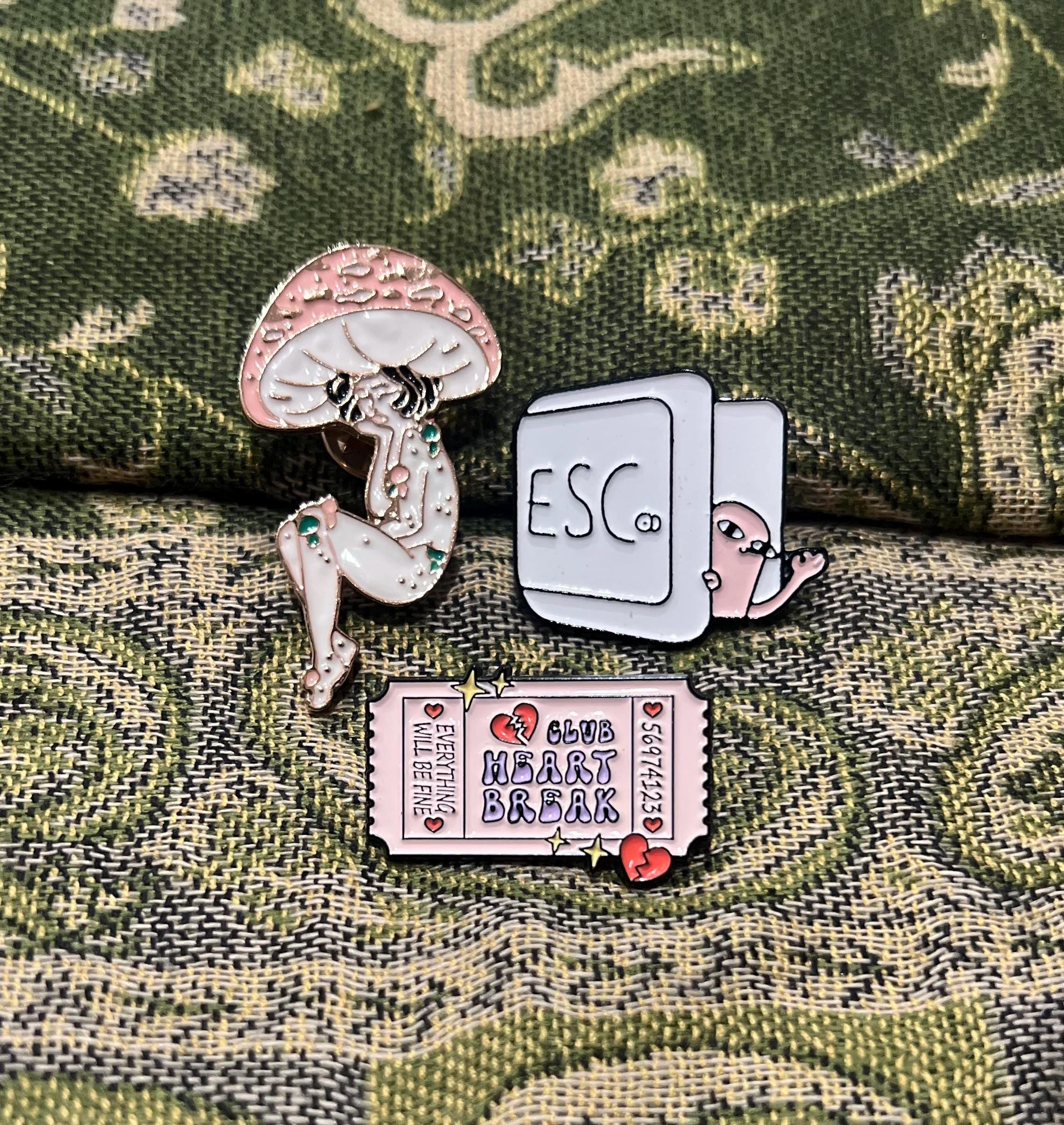 May include: Three enamel pins: a pink mushroom with a human figure inside, a white computer key with the word 'ESC' on it and a pink creature peeking out, and a pink ticket with the words 'Club Heart Break' and 'Everything will be fine'.