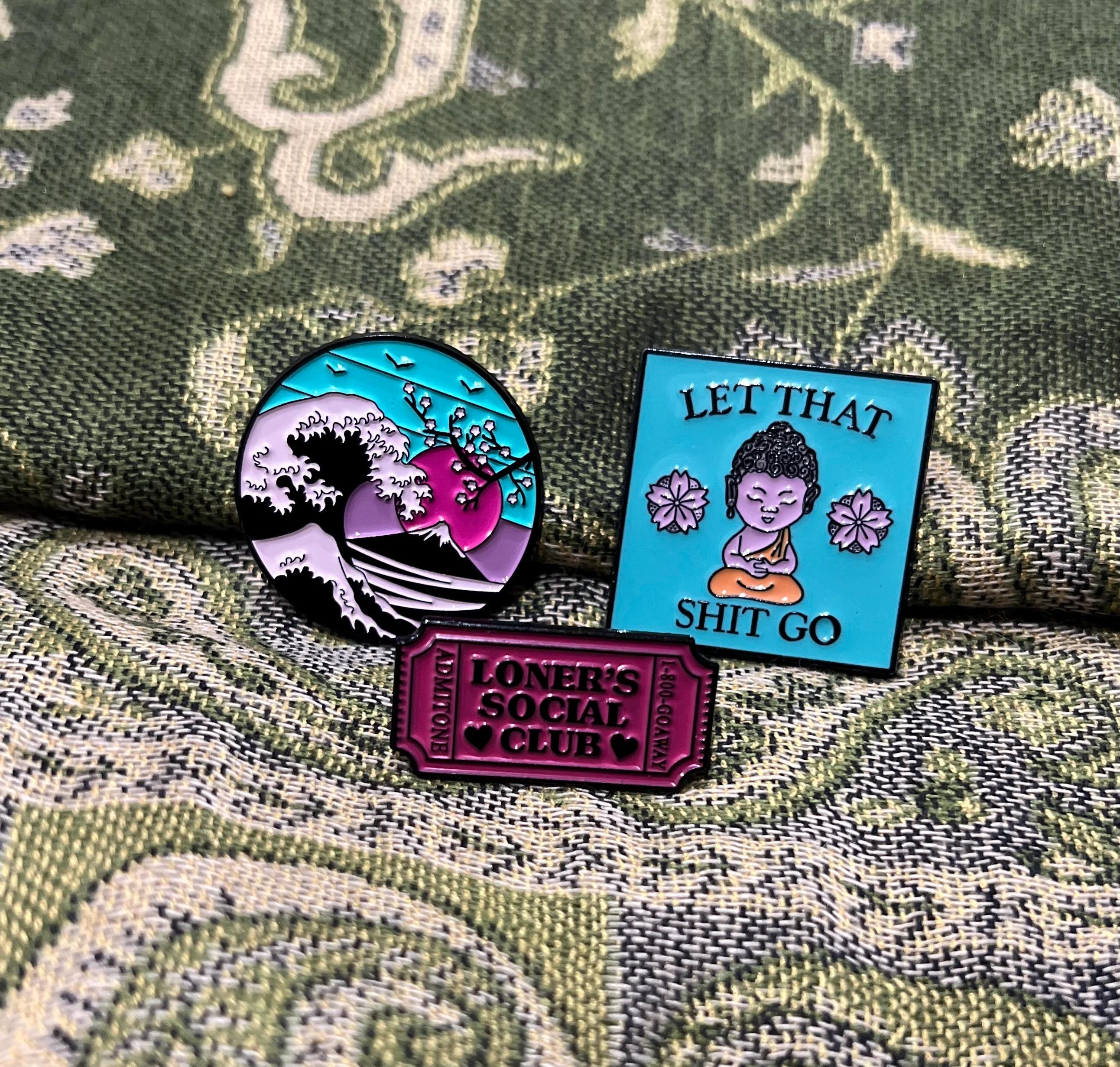 May include: Three enamel pins. One pin is round and blue with a pink and white wave and a pink sun. Another pin is square and blue with the text 'Let That Shit Go' and a purple Buddha sitting on a yellow cushion. The third pin is a ticket shape and purple with the text 'Loners Social Club'.