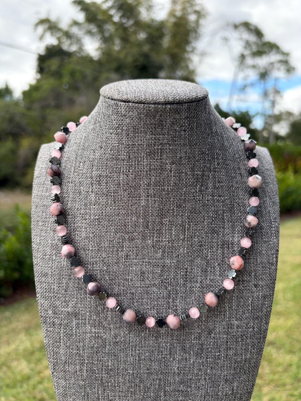 May include: A beaded necklace featuring alternating round pink mottled stone beads and small, dark metallic flower-shaped spacer beads. The beads are strung in a repeating pattern, creating a circular design that rests on a gray fabric display form.